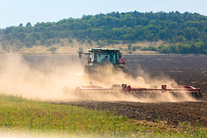 Modern tractor works on a sunlit field, creating dust clouds against a green fo, an Industrial Photo by Ales Munt