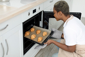 Smiling millennial african american woman chef taking out croissants from oven, a Person Photo by Prostock-Studio