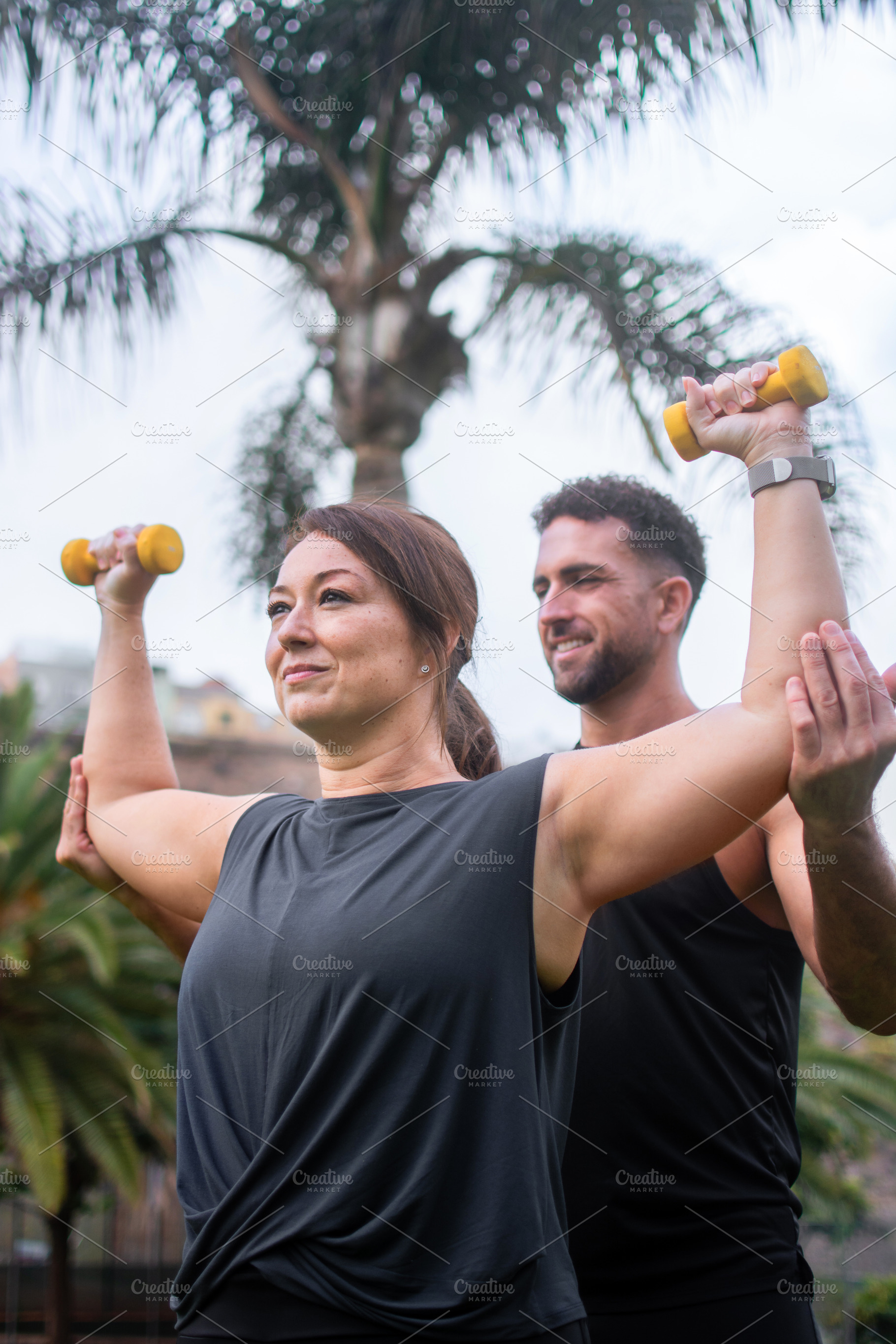Male personal trainer assisting woman with dumbbell exercise outdoors, a Photo by etorres69