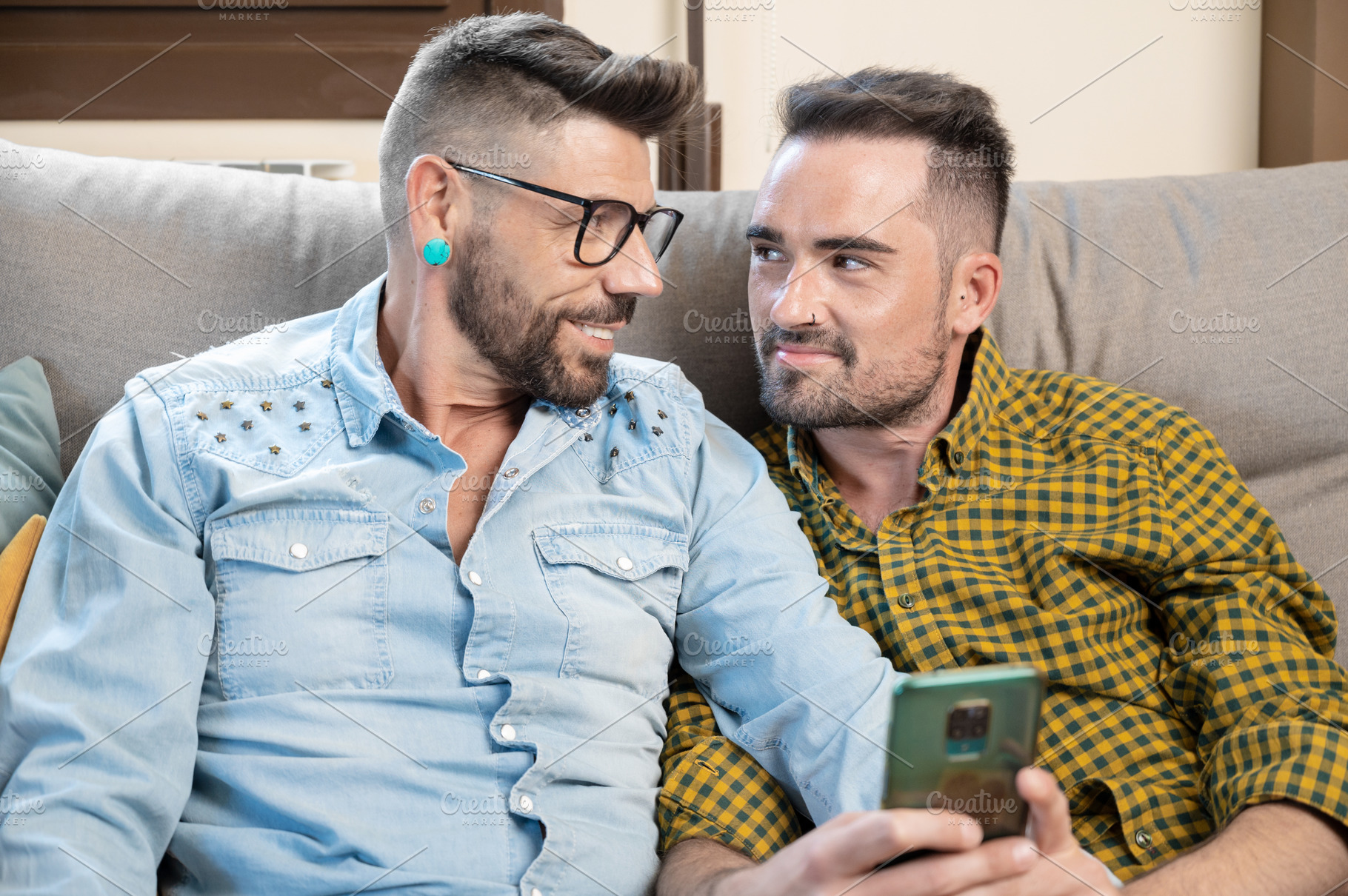 Cheerful gay couple sitting on the couch hugging and looking at the mobile phon, a Person Photo by david.herraez.calzad
