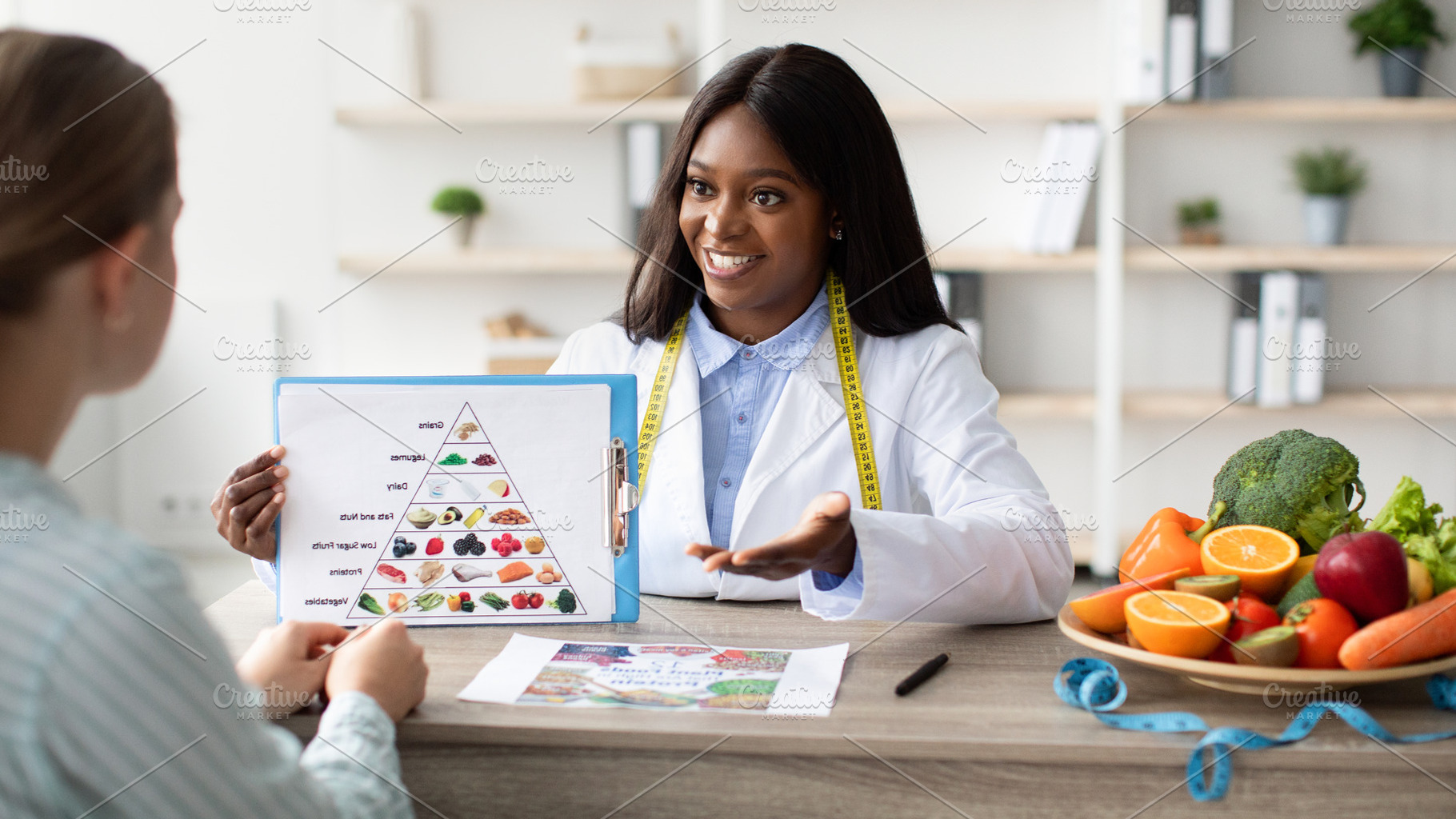 Young black female dietologist showing patient eating food pyramid, having cons, a Person Photo by Prostock-Studio