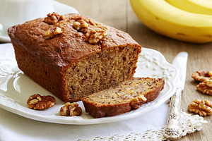 Banana walnut bread loaf, sliced on a white plate with fresh bananas and a knif, a Photo by Studio ANNA