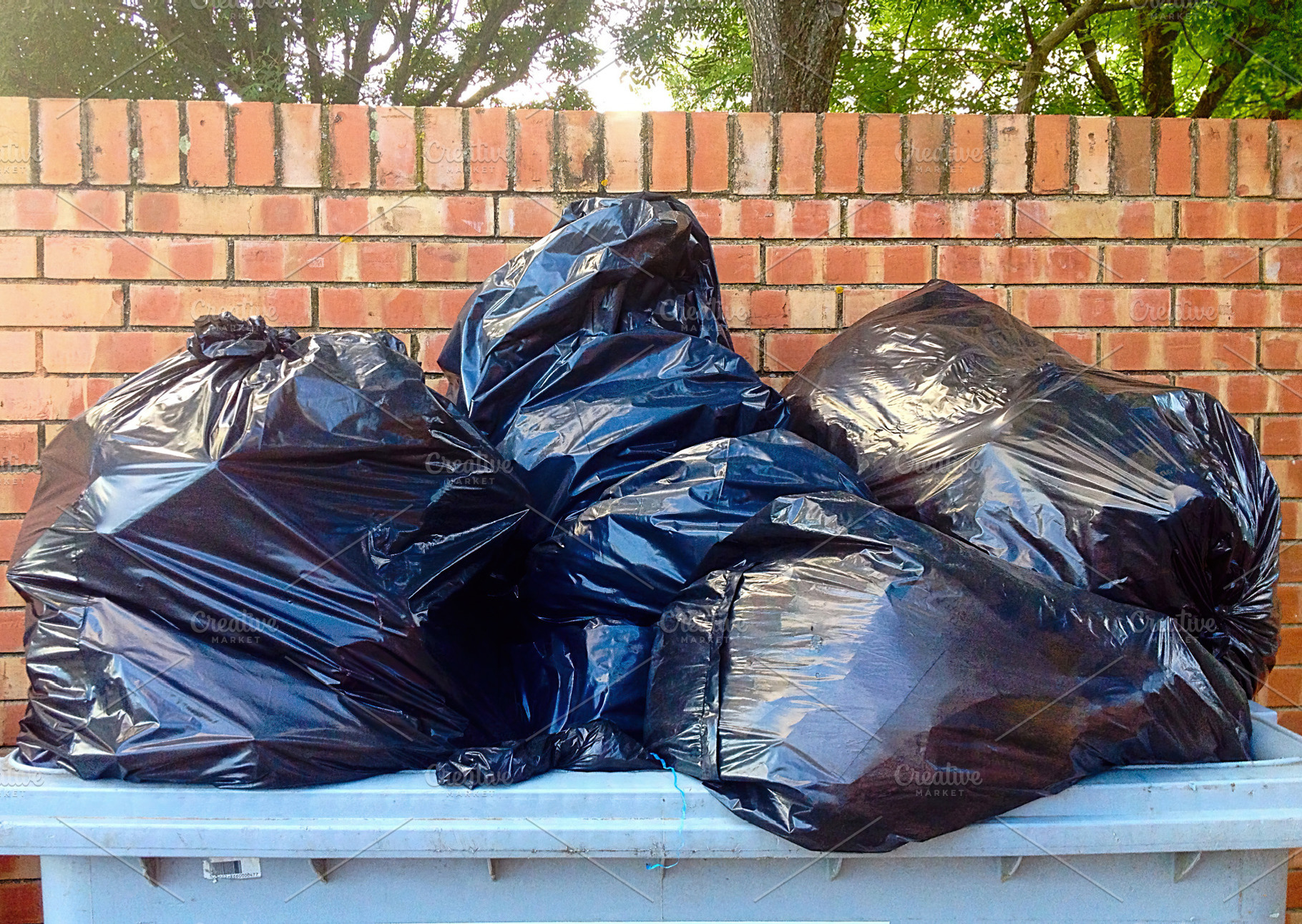 Overflowing garbage bin with black plastic bags near brick wall, an Industrial Photo by Antonio Gravante Market