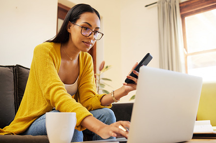 Woman working remote while typing on her laptop and holding her smartphone sitting on a sofa in a bright living room. One focused young hispanic female with glasses at home using modern technology, a Photo by Peopleimages.com