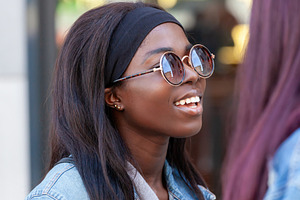 Young black woman wearing sunglasses smiling and chatting with a friend, an Abstract Photo by Antonio Gravante Market