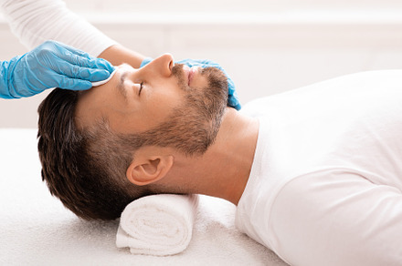 Cosmetologist cleansing bearded man face with toner before treatment, a Photo by Prostock-Studio