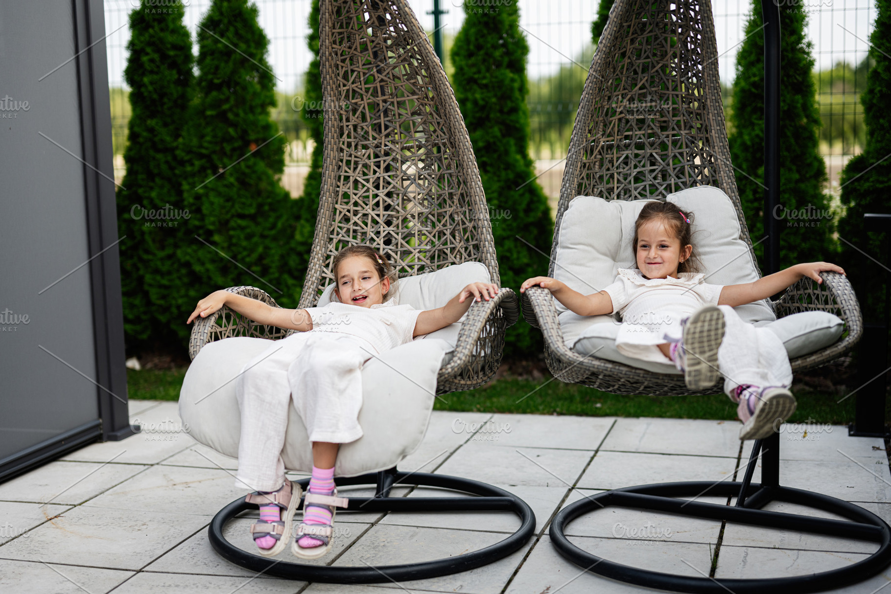 Two young girls enjoying relaxation on modern outdoor wicker egg-shaped chairs, a Photo by AS photostudio