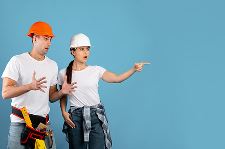 Shocked Young Couple In Hard Hats Pointing Aside At Copy Space, a Background Photo by Prostock-Studio