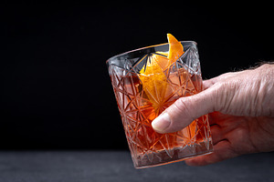 Hand holding classic old fashioned cocktail with orange garnish on dark backgro, a Food & Drink Photo by etorres69