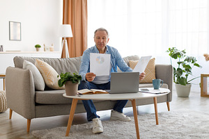 Man holding paper reading report working on laptop at home, a Photo by Prostock-Studio