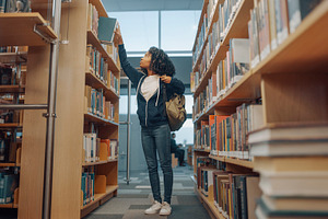 Black teenage student with curly hair reaching for a book on an, a School & Education Photo by DanFLCreativo