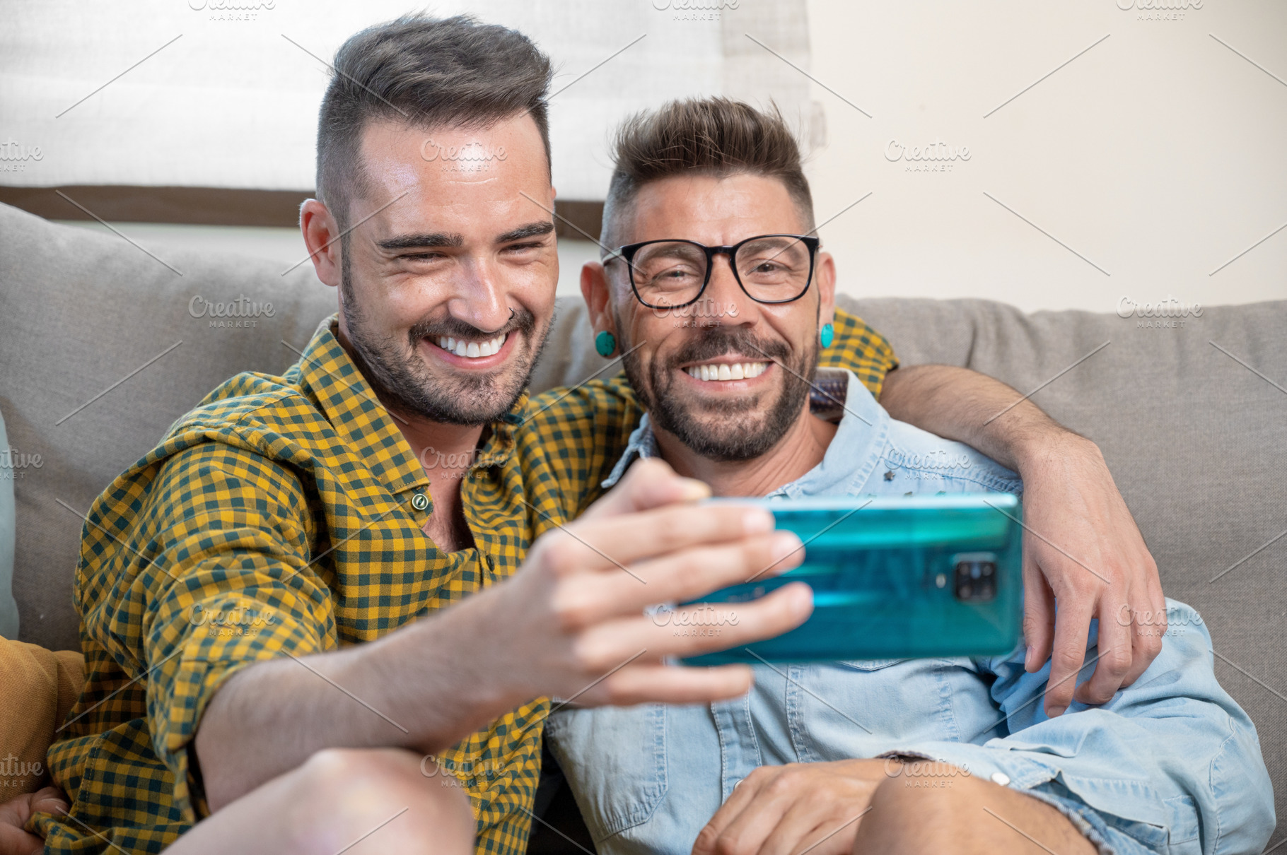 Cheerful gay couple sitting on the couch hugging and looking at the mobile phon, a Person Photo by david.herraez.calzad