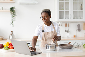Smiling millennial african american female in glasses, apron cooking, watching, a Person Photo by Prostock-Studio