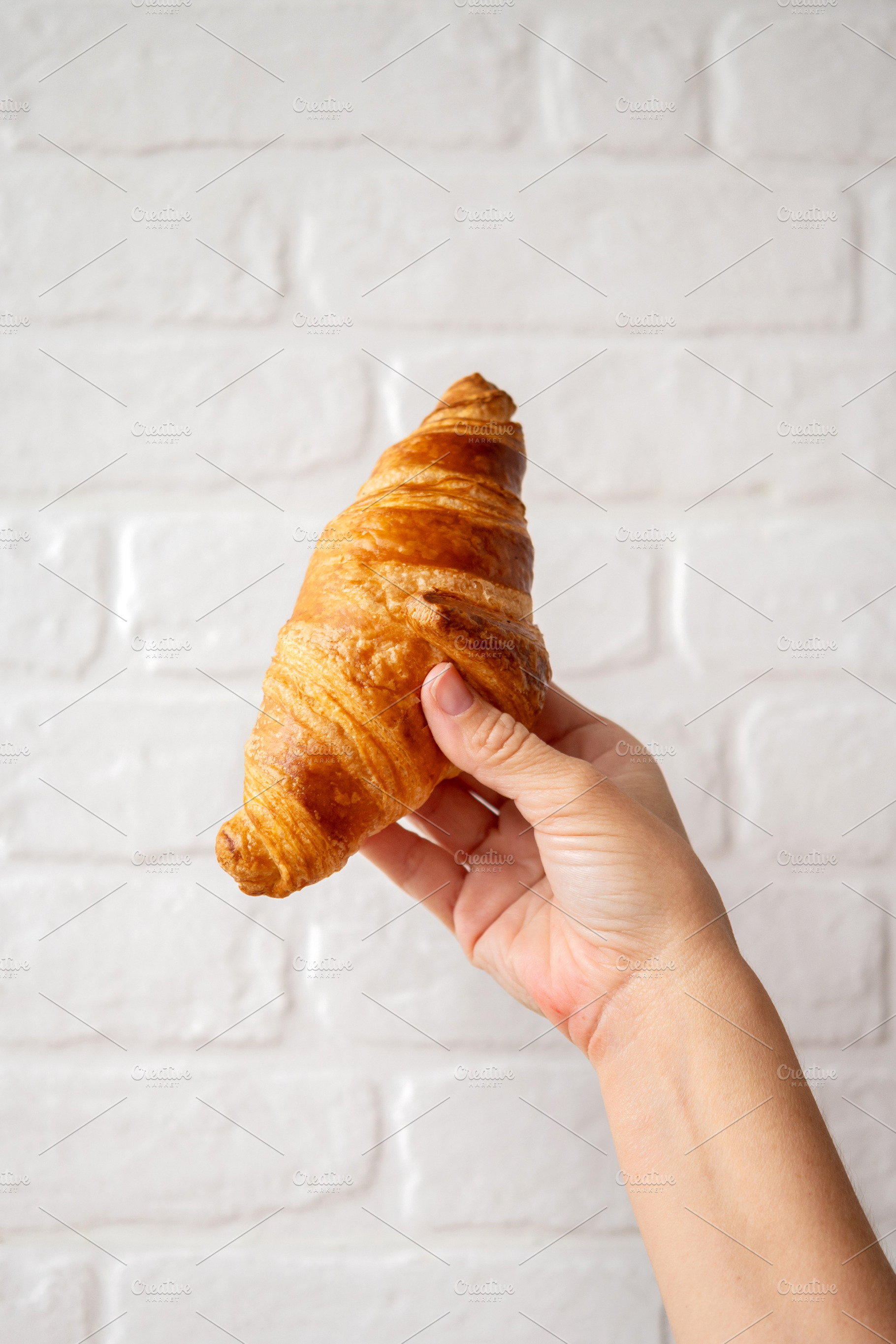 Hand Holding a Fresh Butter Croissant Against a White Brick Wall, a Food & Drink Photo by etorres69