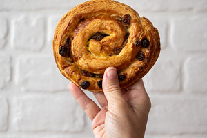Hand holding pain aux raisins against white brick wall, a Food & Drink Photo by etorres69