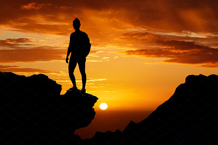 Sunset, hiking and silhouette of a man on top of the mountain during an adventure journey in africa. Freedom, orange sky and guy standing on a cliff while trekking in nature on a summer evening., a Photo by Peopleimages.com