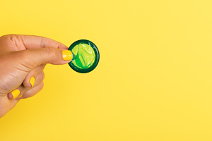 Hand with yellow nails holding a green rolled condom against a, a Health & Medical Photo by Ales Munt