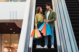 Couple Shopping Holding Bags Standing On Moving Stairs In Mall, a Photo by Prostock-Studio