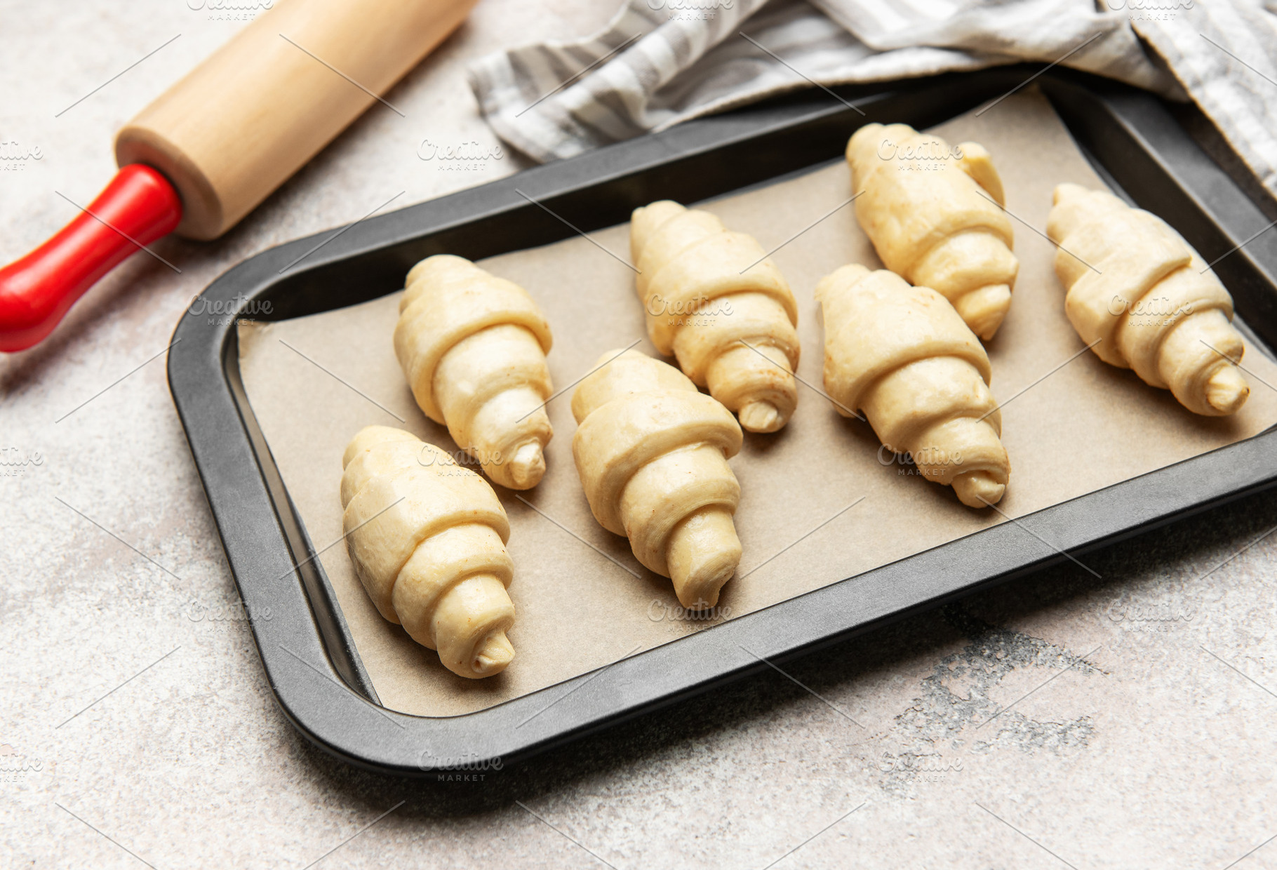 Raw croissants baking on tray with rolling pin and kitchen towel, a Food & Drink Photo by Olena Rudo