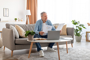 Man holding documents reading report working on laptop at home, a Photo by Prostock-Studio