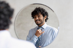 Handsome indian businessman putting on necktie and looking at his reflection in, a Person Photo by Prostock-Studio
