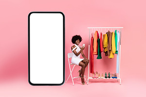 African American Lady Using Cellphone Shopping Choosing Clothes, Pink Backgroun, a Photo by Prostock-Studio