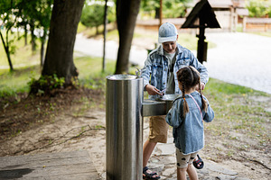 Two children using an outdoor water fountain in a natural park setting, a Person Photo by AS photostudio