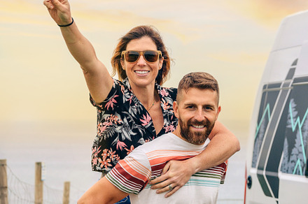 Couple looking camera having fun piggybacking next to their camper van, a Holiday Photo by davidpereiras