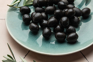 Black olives served on ceramic plate with olive branches on tile, a Food & Drink Photo by Olena Rudo