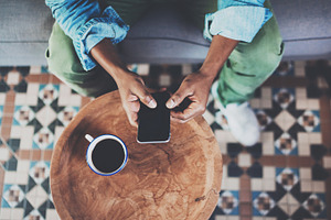 Top view of african man using smartphone while sitting on sofa at home.Concept people working with mobile gadget.Black coffee wooden table.Blurred background., a Business Photo by SFCRCH MOCKUPS