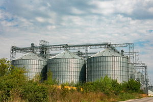 Large metal grain silos under a cloudy sky, surrounded by green vegetation, ill, an Industrial Photo by Ales Munt