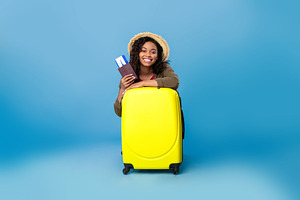 Happy black lady in casual wear sitting behind suitcase, holding passport and f, a Photo by Prostock-Studio