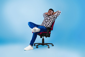 Handsome happy young black man relaxing in chair over blue studio background, f, a Photo by Prostock-Studio