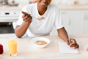 Smiling millennial black female eating porridge at table with cookies, juice an, a Person Photo by Prostock-Studio