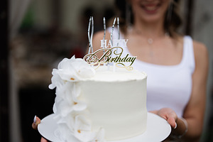 Elegant white birthday cake with decorations held by a smiling woman, a Photo by AS photostudio