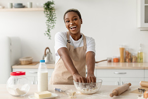 Happy laughing young black female in apron making dough, enjoy cooking pie in c, a Photo by Prostock-Studio