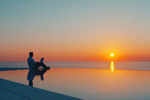 Man working on laptop by infinity pool at sunset over ocean, a Photo by AlyaAnd