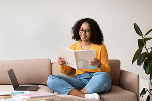 Cheery African American woman sitting on couch with laptop and book, doing home, a Photo by Prostock-Studio