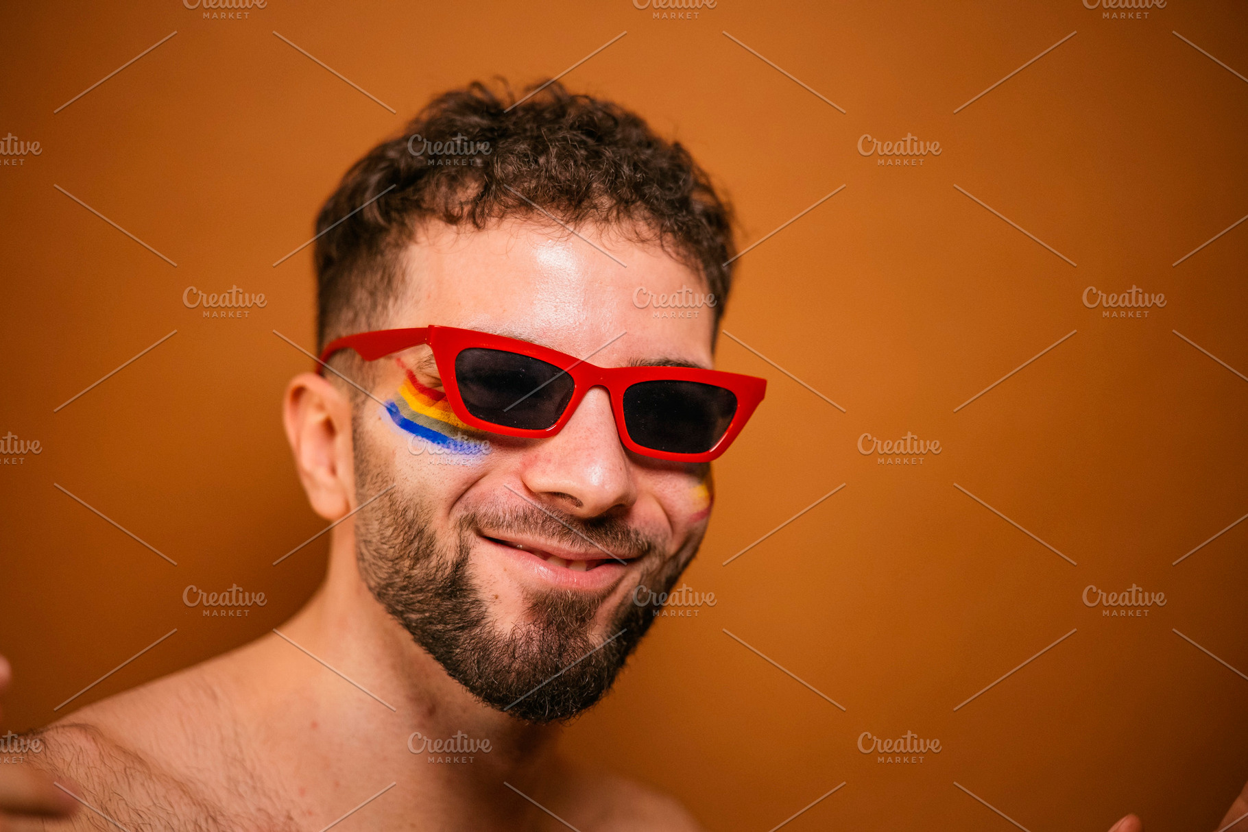 Young man smiling wearing red sunglasses with rainbow paint, a Photo by karrastock