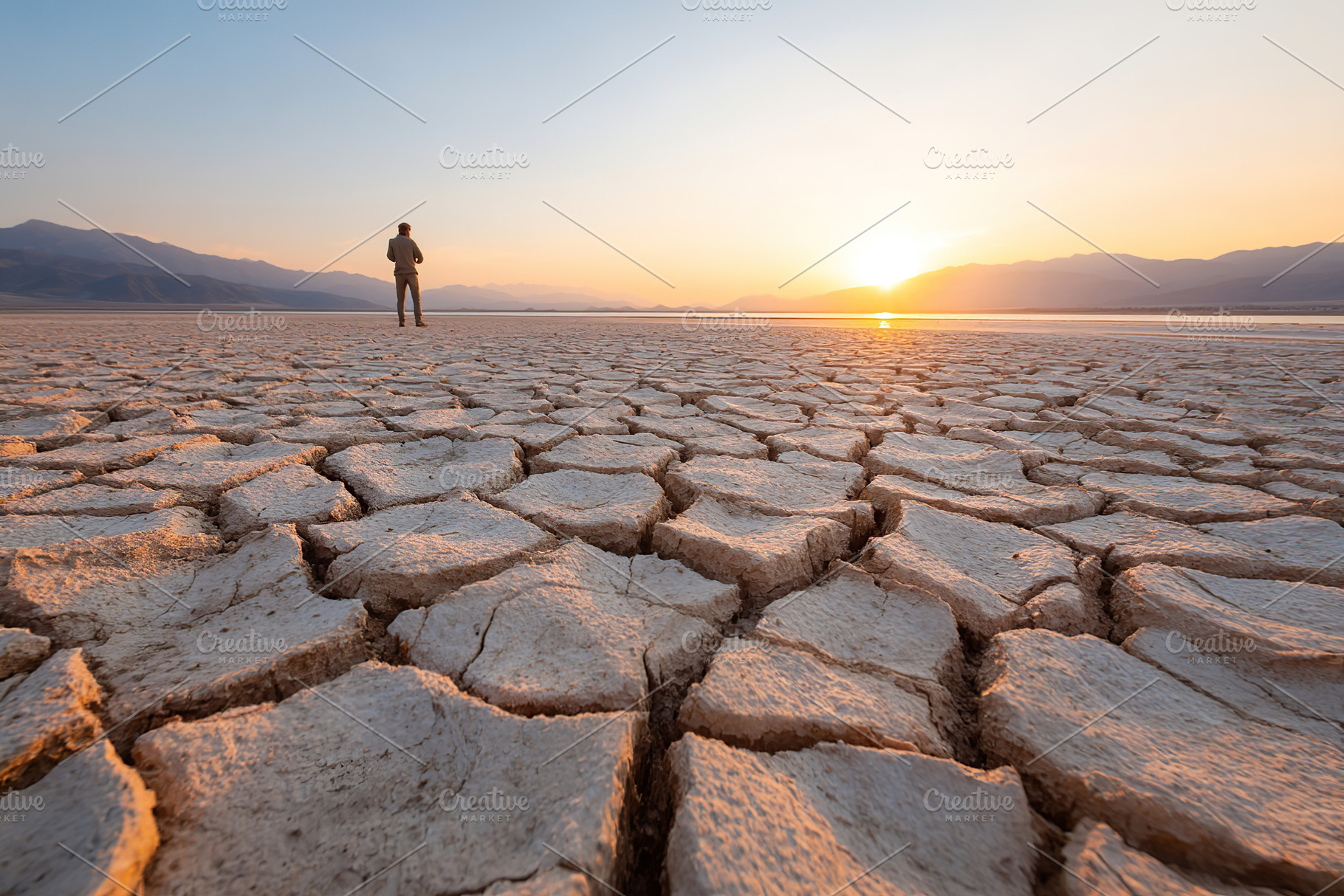 Cracked earth landscape at sunset with figure standing in vast desert expanse, a Photo by AlyaAnd
