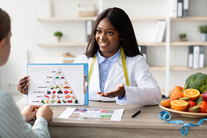 Young black female dietologist showing patient eating food pyramid, having cons, a Person Photo by Prostock-Studio