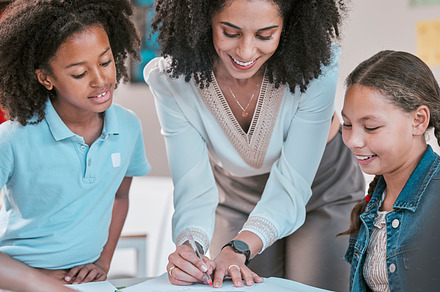 Helping students with homework, female teacher in classroom with children listening and writing in book. Diversity in education, educator reading kids notebook and group learning literature together, a Photo by Peopleimages.com