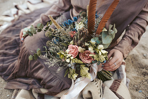 Bride in brown clothes with a rustic bouquet sit on the sand artwork country