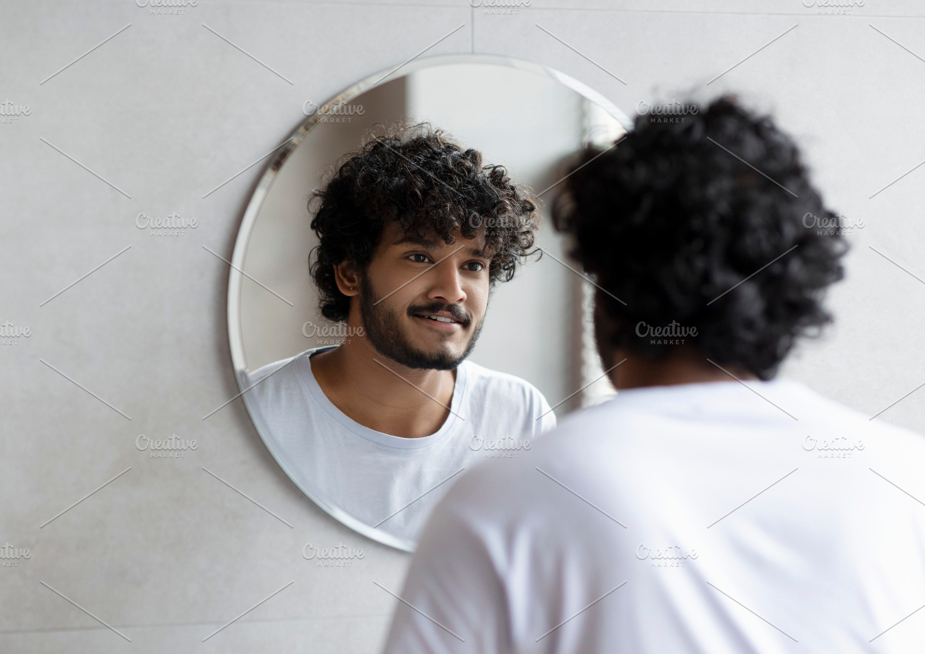 Handsome indian guy looking at his reflection in the mirror, standing in modern, a Person Photo by Prostock-Studio