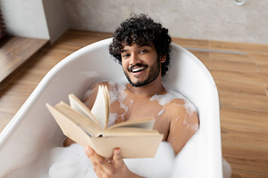 Happy indian man lying in foamy bath and reading book in relaxing atmosphere in, a Person Photo by Prostock-Studio