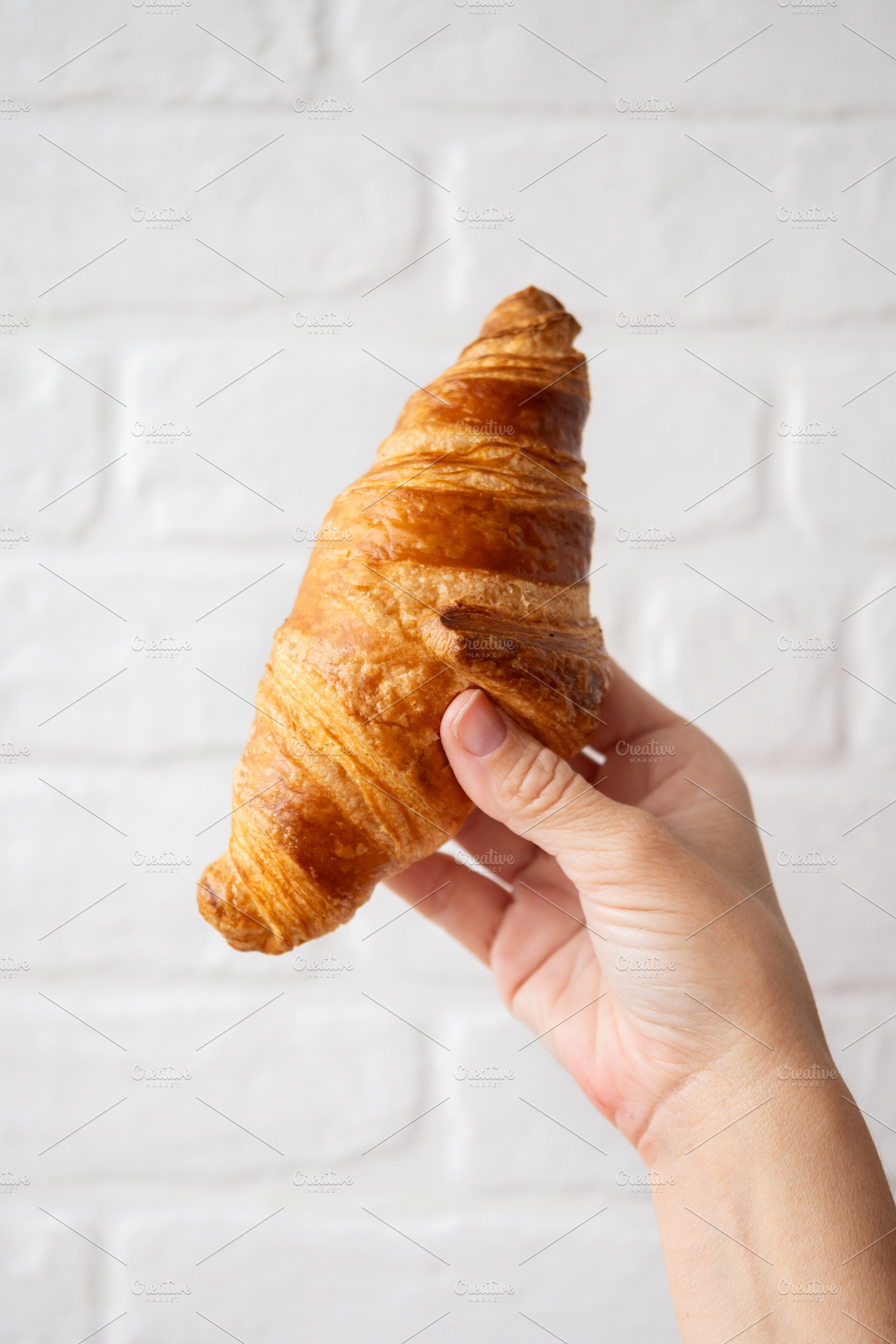 Hand Holding Fresh Croissant Against White Brick Background, a Food & Drink Photo by etorres69