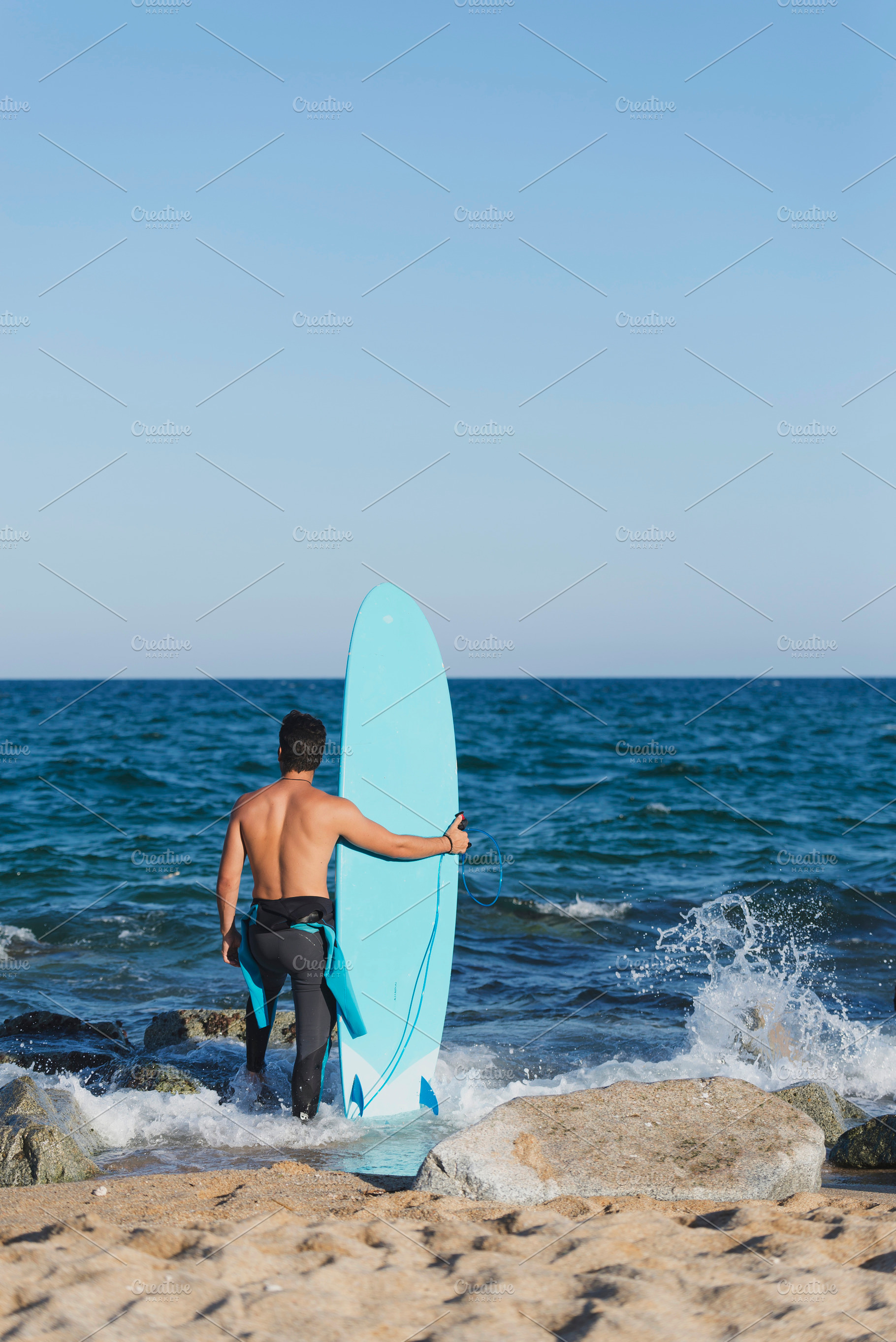 Back view of young surfer standing barefooted on sandy shore,, a Holiday Photo by 2Design