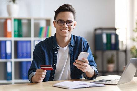 Young Man With Smartphone And Credit Card Sitting At Desk In Office