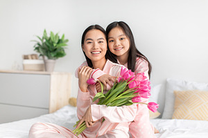 Cheerful chinese young girl hug millennial lady in pink pajama with tulips bouq, a Photo by Prostock-Studio