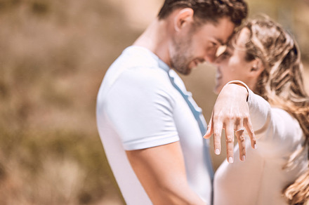 Closeup of happy female hand engaged and romantic couple in love after outdoors marriage proposal. Woman wearing engagement ring with smiling, affectionate lovers hugging and embracing in background., a Photo by Peopleimages.com